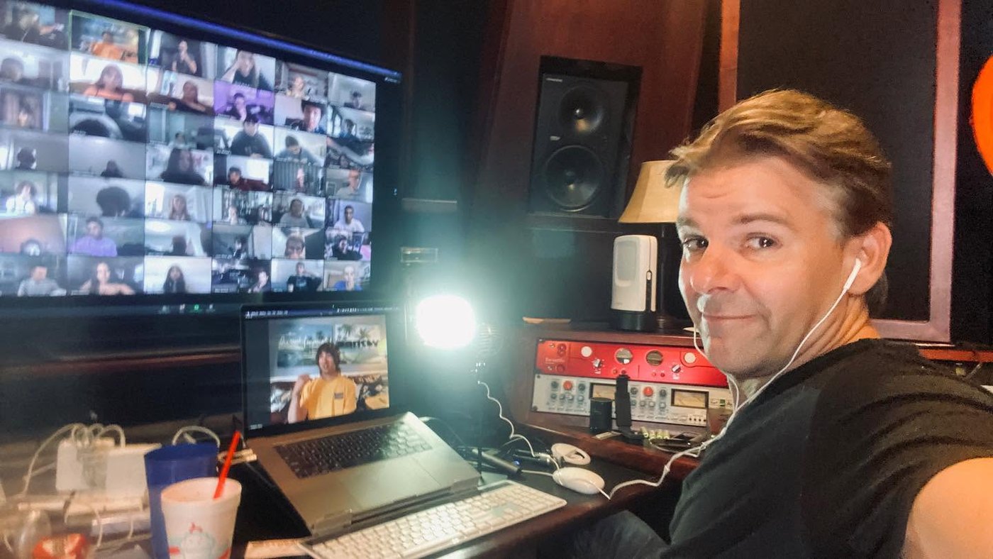 Nathan Adam in a studio control room facing a video call grid of dozens of students on a large monitor