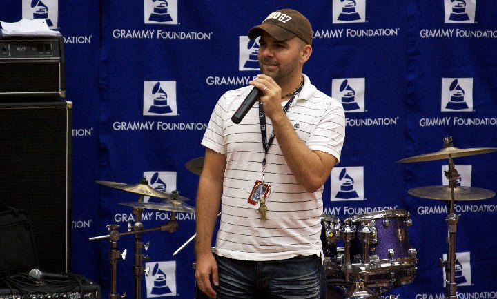 Nathan Adam speaking with a microphone in front of a GRAMMY Foundation backdrop