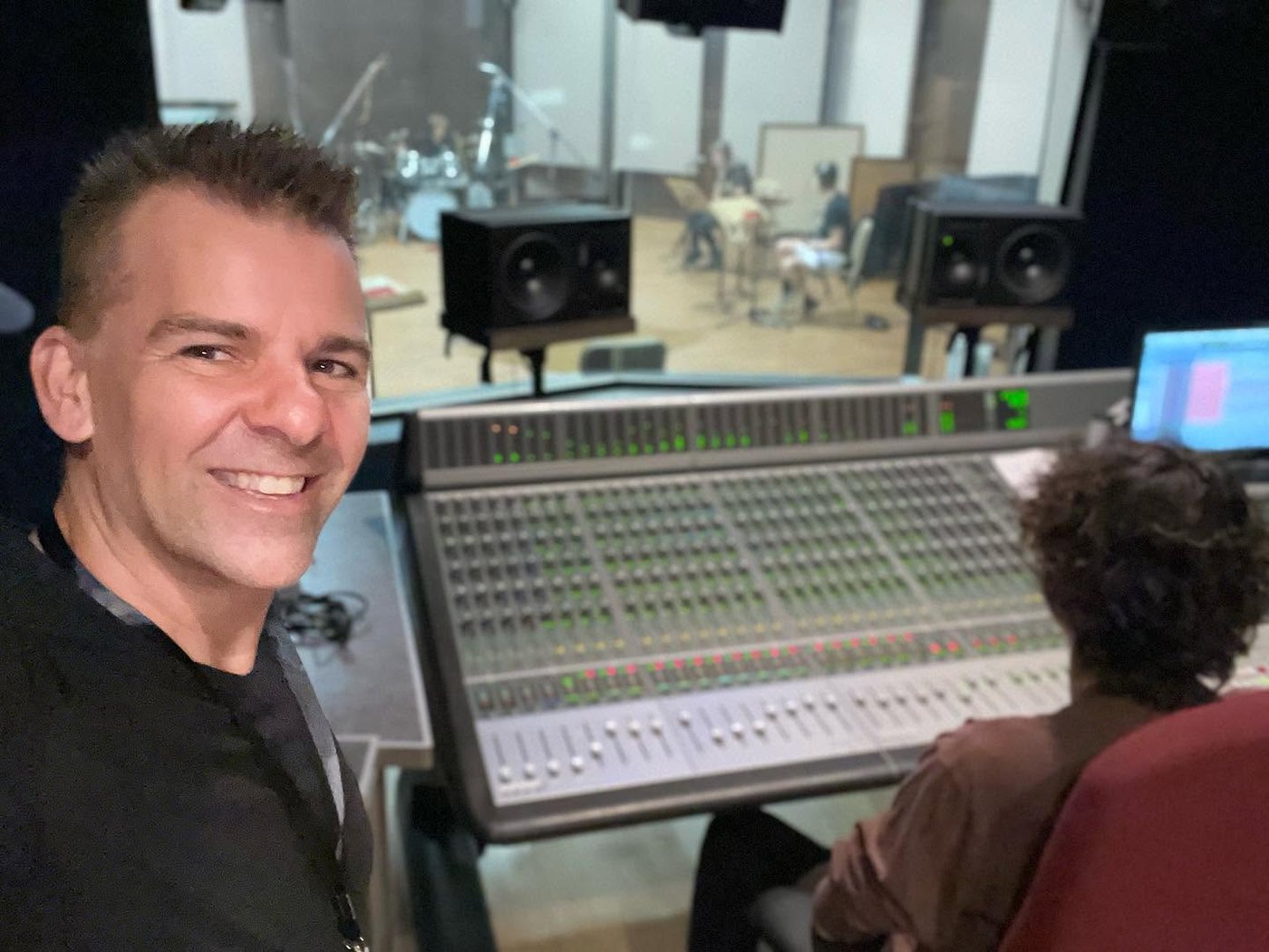 Nathan Adam at a large mixing console with a band tracking in the live room beyond the glass